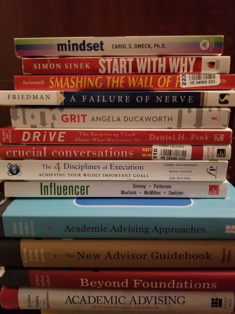 Personal photo of the 13 book bindings on a stack of my current (and growing) future readings. The book titles are Mindset, Start with Why, Smashing the Wall of Fear, A Failure of Nerve, Grit, Drive, Crucial Conversations, the 4 Disciplines of Execution, Influencer, Academic Advising Approaches, The New Advisor Guidebook, Beyond Foundations, and Academic Advising a Comprehensive Handbook