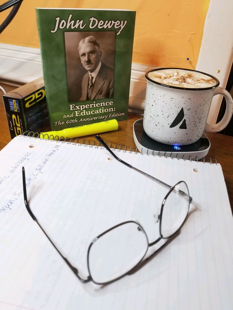 Image of authors workspace. A pair of reading glasses is resting on a spiral notebook with a background of a full box of pens, a highlighter, John Dewey's book, and a cup of cream topped coffee sits on a coffee warmer.