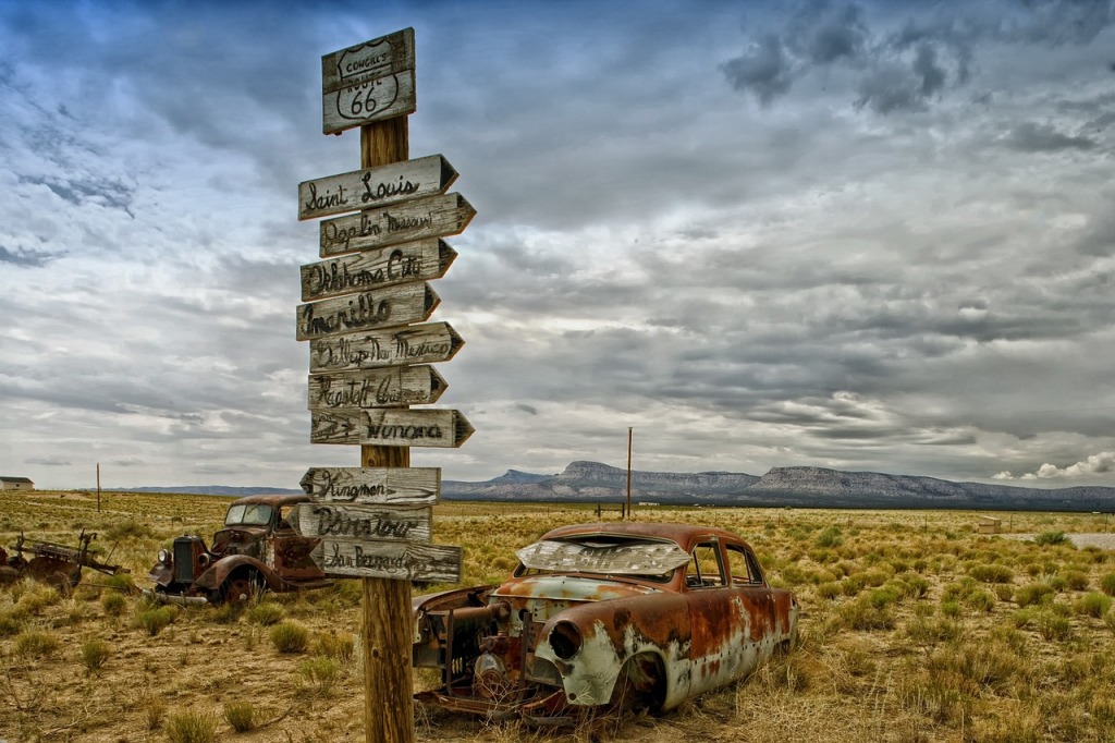 A wooden signpost is covered with hand written white wooden arrows to towns on Route 66 with rusty cars in the background of a scenic cloudy desert landscape with mountain range far in the distance.