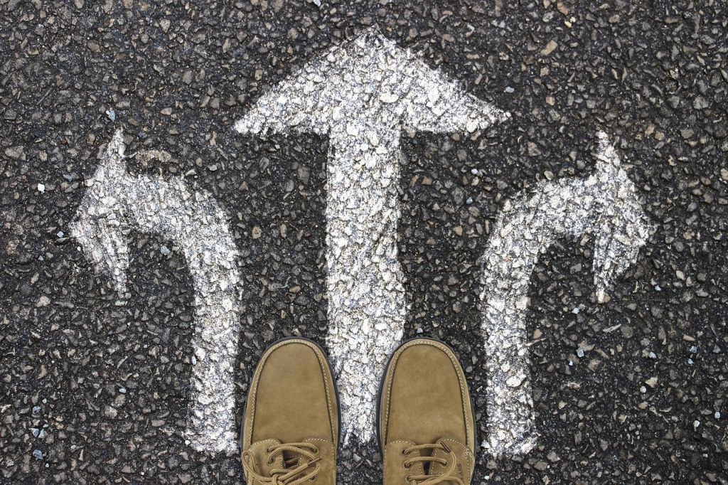 Three arrows (left, straight, right) are painted on asphalt with shoed feet standing at the base of each.